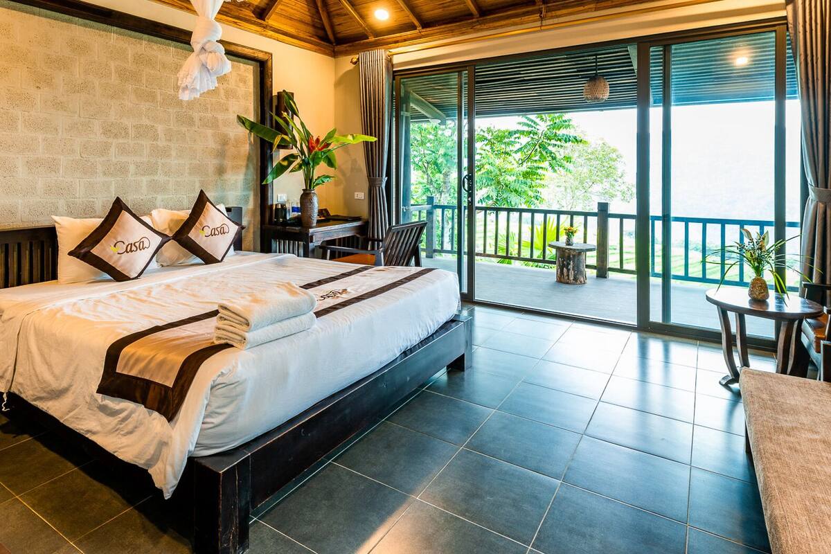 A hotel room with a bed, a nightstand, a potted plant, and a view of the outdoors.