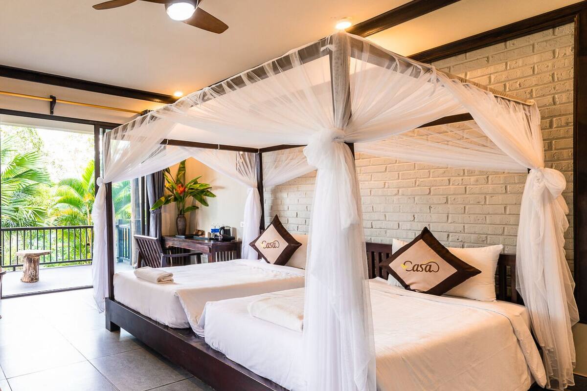A double bed with a canopy, two pillows with the word "Casa" on them, a ceiling fan, and a brick wall.