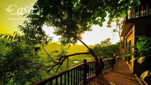 A wooden pathway leading to a building with a balcony, surrounded by lush greenery and a view of distant hills. - Puluong Casa Resort (Pu Luong)