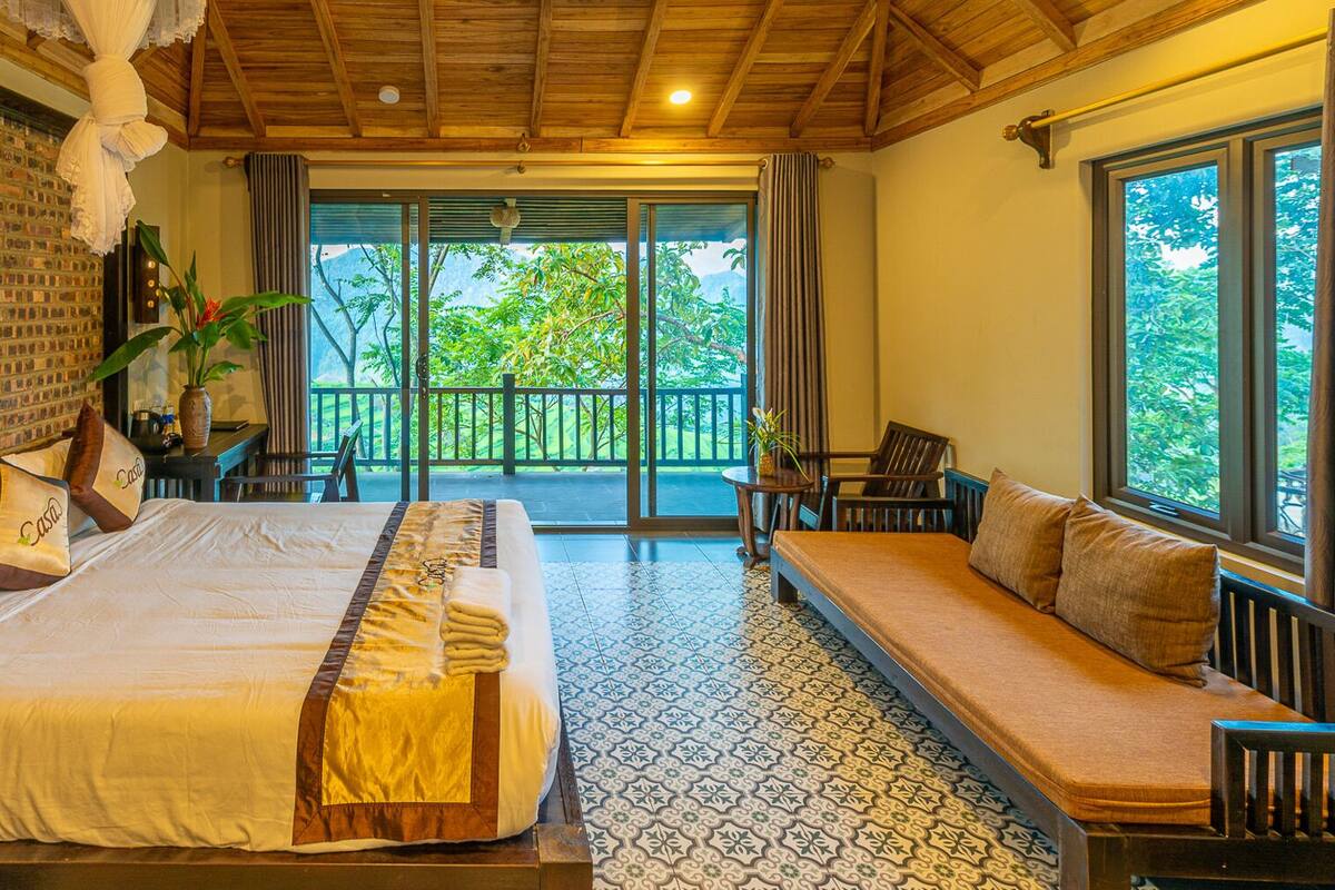 A hotel room with a large bed, a sofa, a balcony with a view of greenery, and a brick wall.