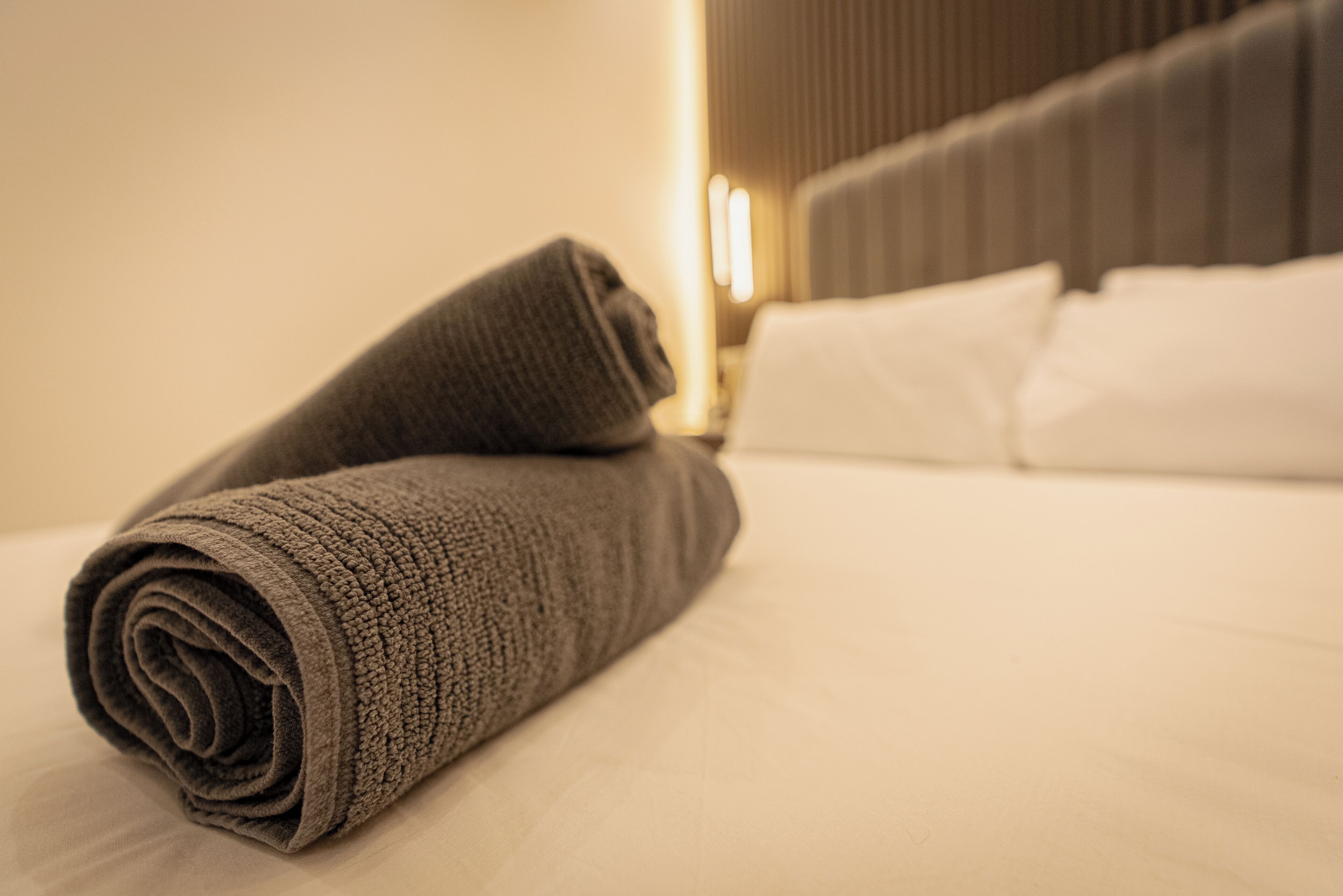 Toalha cinza dobrada sobre uma cama com lençóis brancos, num quarto com cabeceira moderna e luz quente ao fundo.
