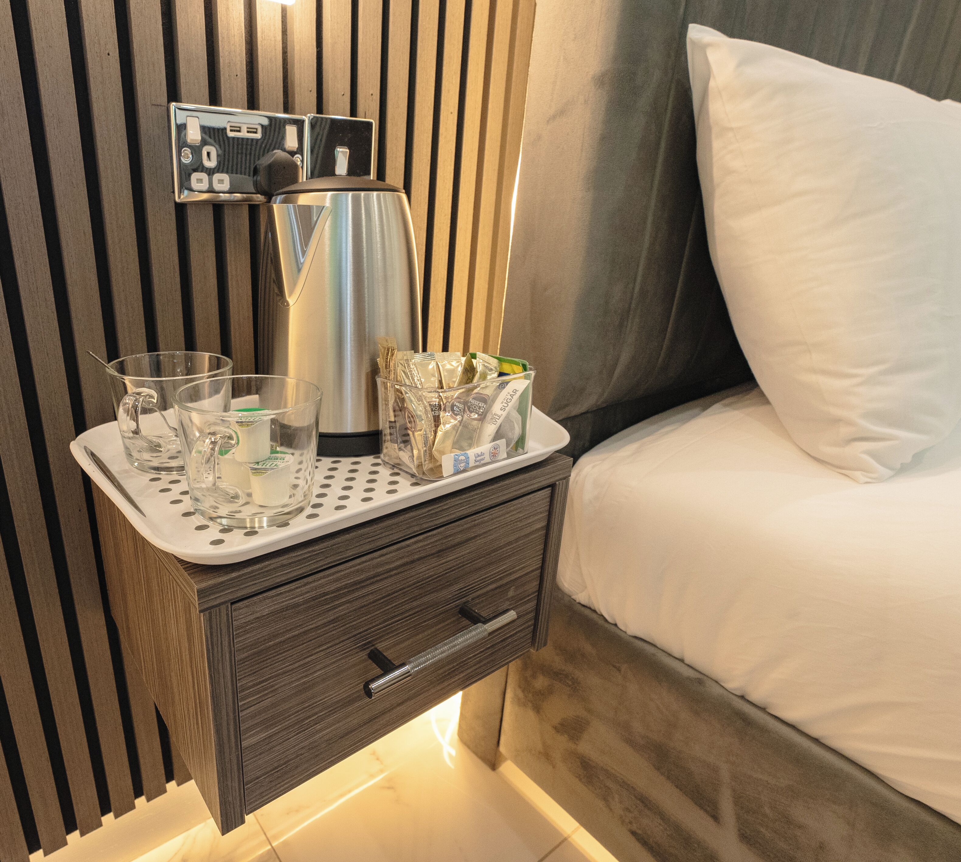 A neatly made bed with a pillow, a bedside table with a kettle, glasses, and a tray with packets.