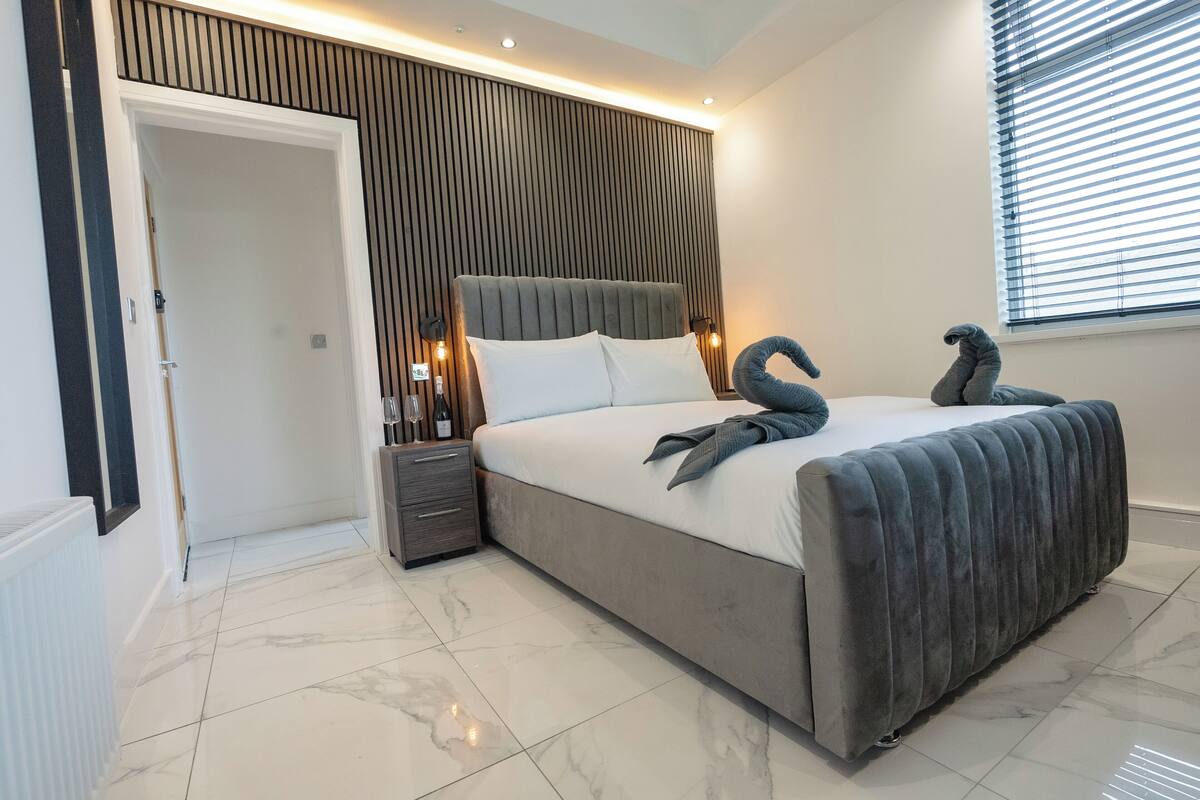 Une chambre moderne avec un grand lit, une table de chevet, une lampe fixée au mur et une fenêtre avec des stores.