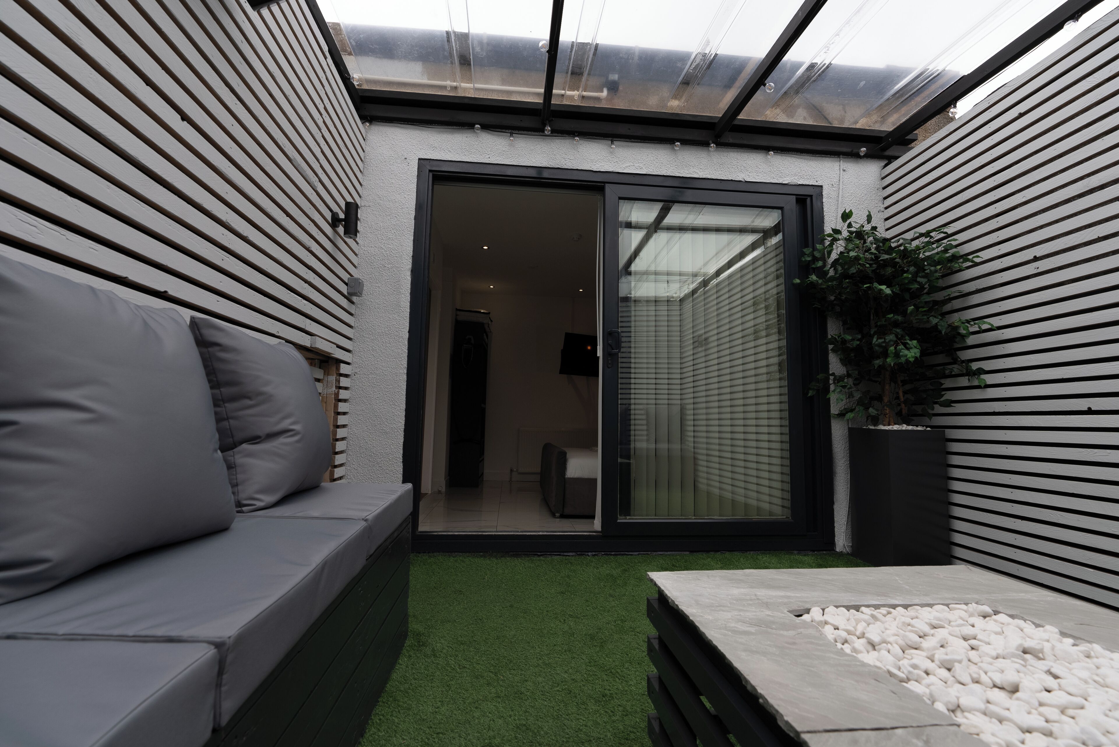 Un patio moderno al aire libre con puerta corrediza de vidrio que da a una habitación, un sofá gris y un pequeño jardín con guijarros blancos.