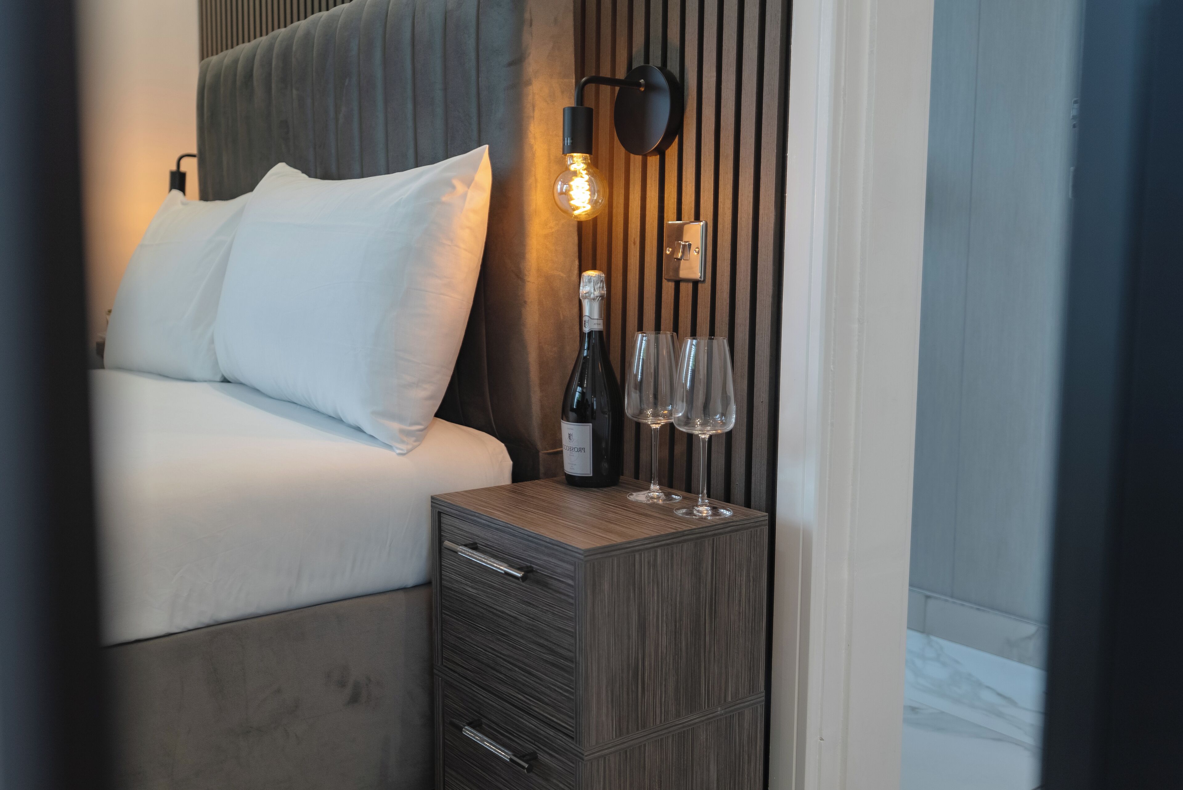 A neatly made bed with white linens, a bedside table with a bottle of wine and two glasses, and a wall-mounted light fixture.