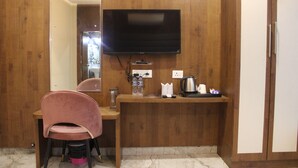 A hotel room with a wooden desk, a pink chair, a flat-screen TV mounted on the wall, and a small coffee station. - Hotel Jsr Ganga (Varanasi)