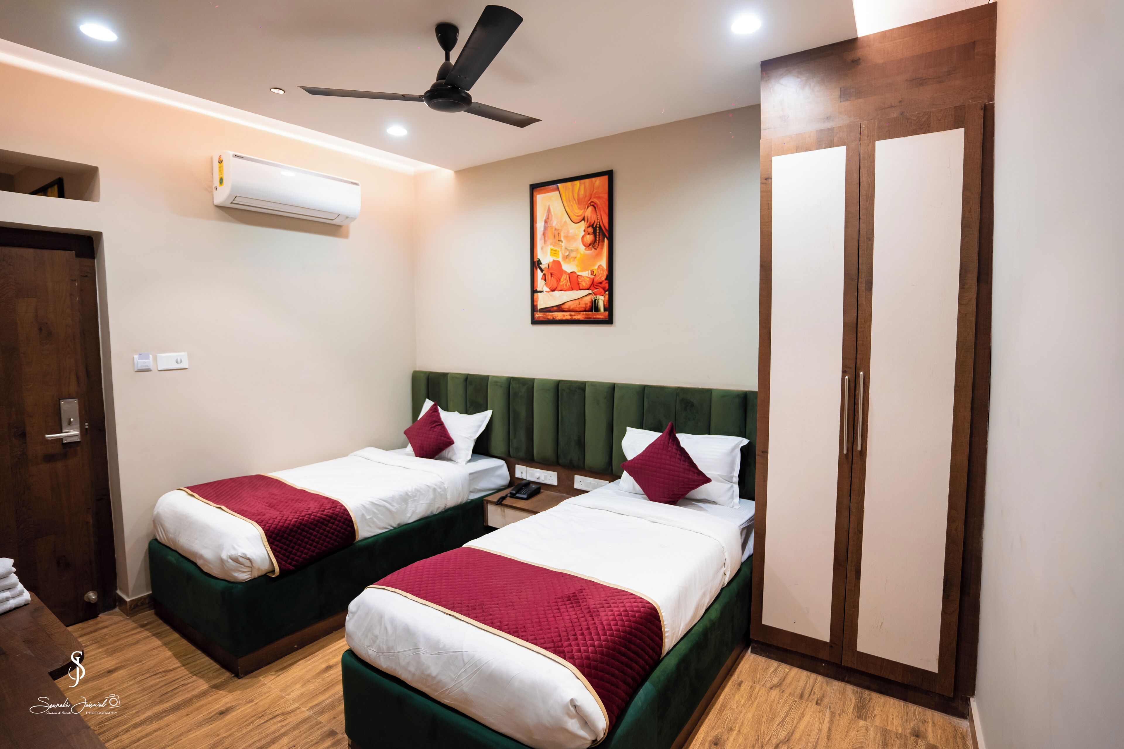 A hotel room with two beds, a ceiling fan, an air conditioning unit, a painting on the wall, and a wardrobe.