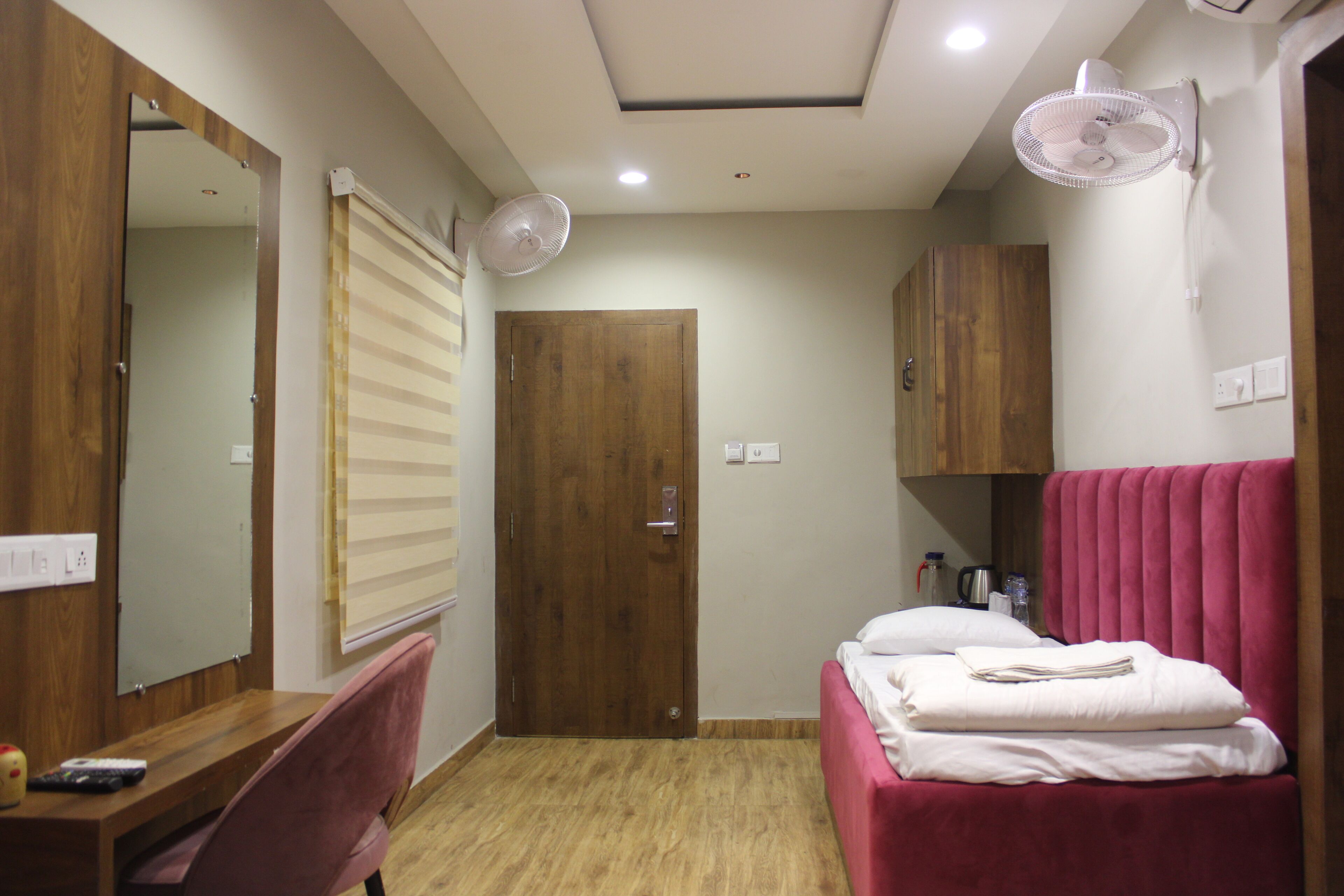 Un dormitorio con puerta de madera, una cama con cabecera rosa, un escritorio con silla y un espejo.