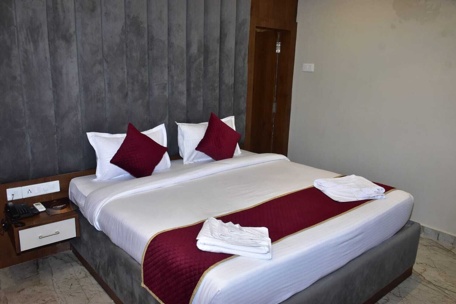 A neatly made bed with white linens and red pillows, a wooden nightstand with a phone and a small drawer, and a marble floor.