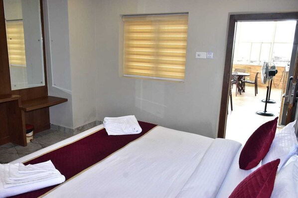 Habitación de hotel con cama, banco de madera, ventana con persianas y vista al exterior.