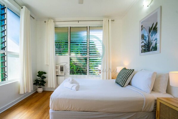Een slaapkamer met een bed, witte gordijnen, een kamerplant en een raam met zonwering.