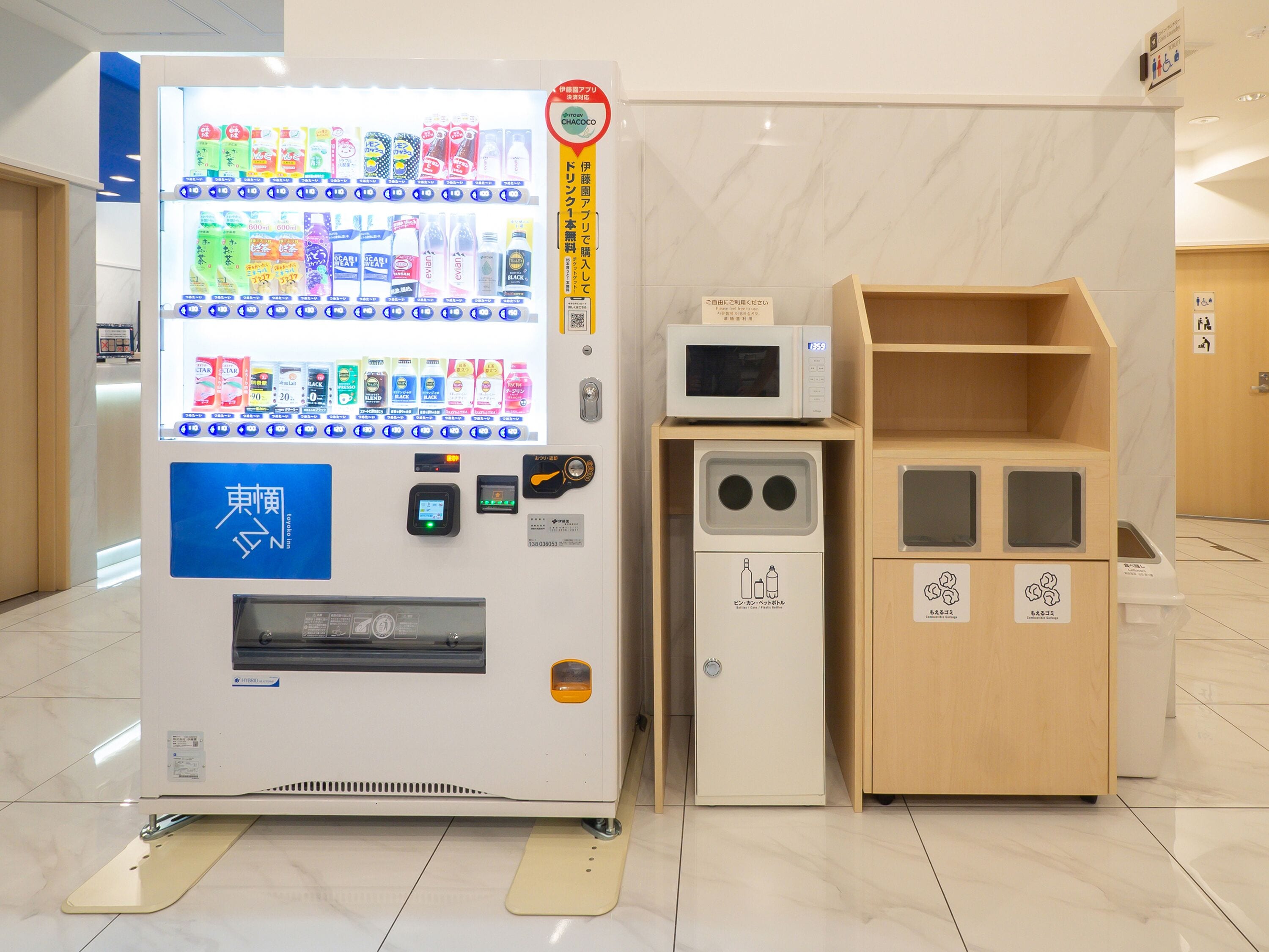 Máquina expendedora con diversas bebidas y aperitivos, un microondas y un pequeño armario de madera con símbolos de reciclaje.