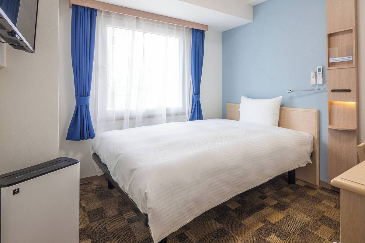 A hotel room with a bed, a TV, a desk, and a window with curtains.