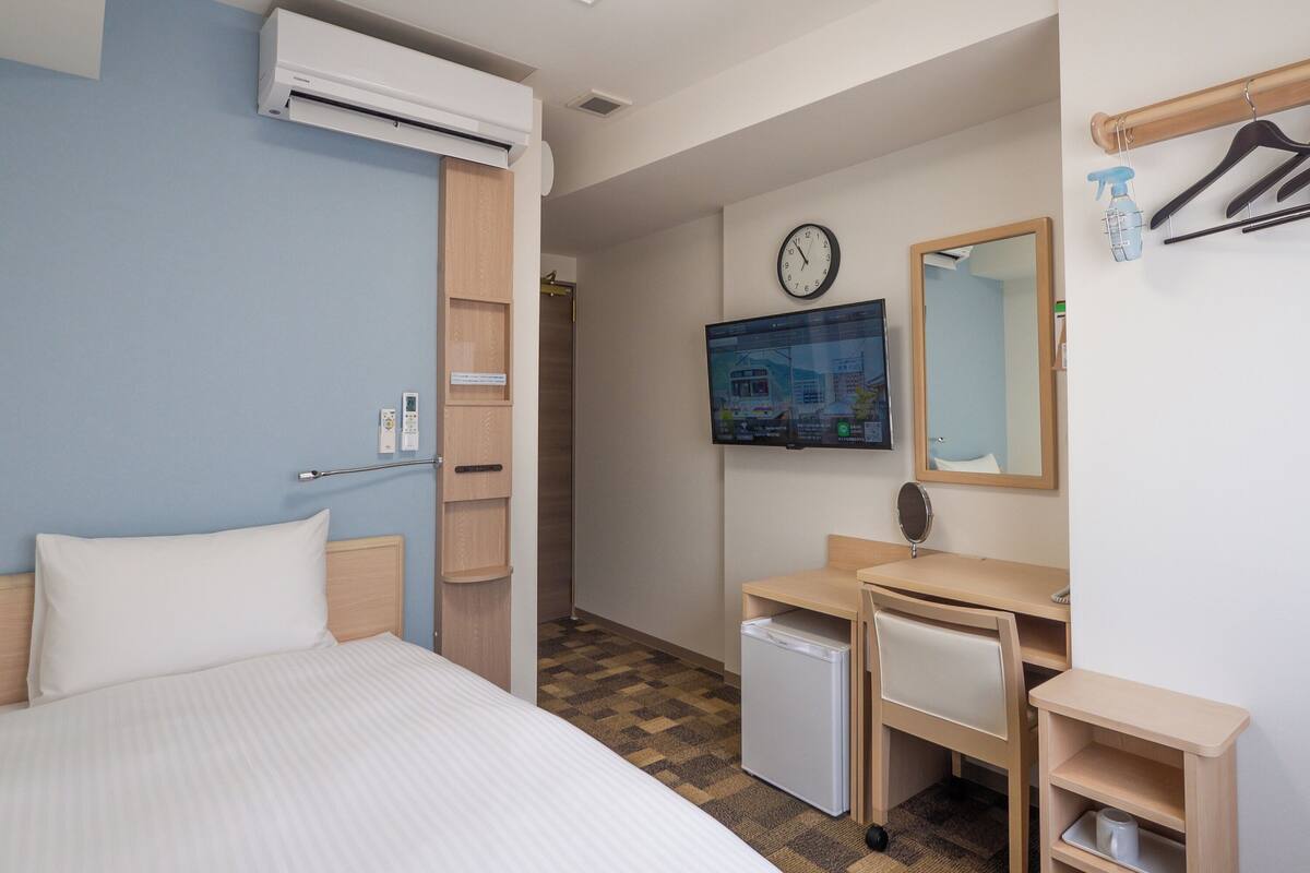 A hotel room with a bed, a desk, a small refrigerator, a clock, and a television.