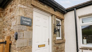 A stone cottage with a white door, a sign reading "Hall Garth Cottage," and a small window with a lantern. - Hall Garth Cottage (Leyburn)