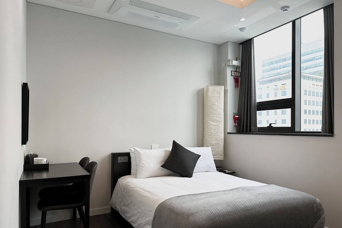 A hotel room with a bed, a desk, a chair, a large window with a city view, and a mounted air conditioner.