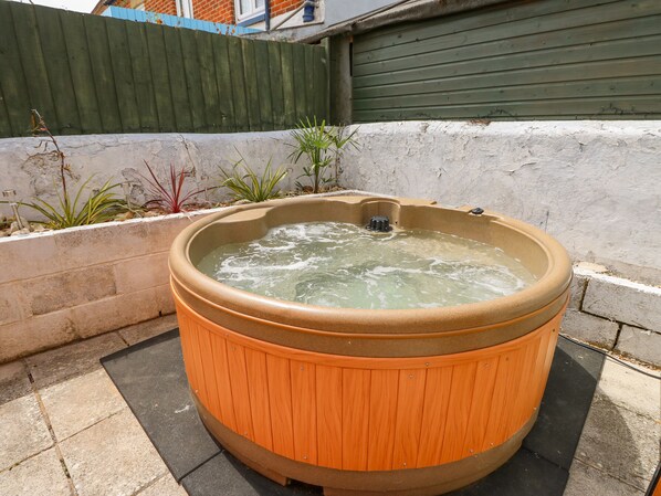 A wooden hot tub in an outdoor setting with a concrete floor and a white wall in the background.