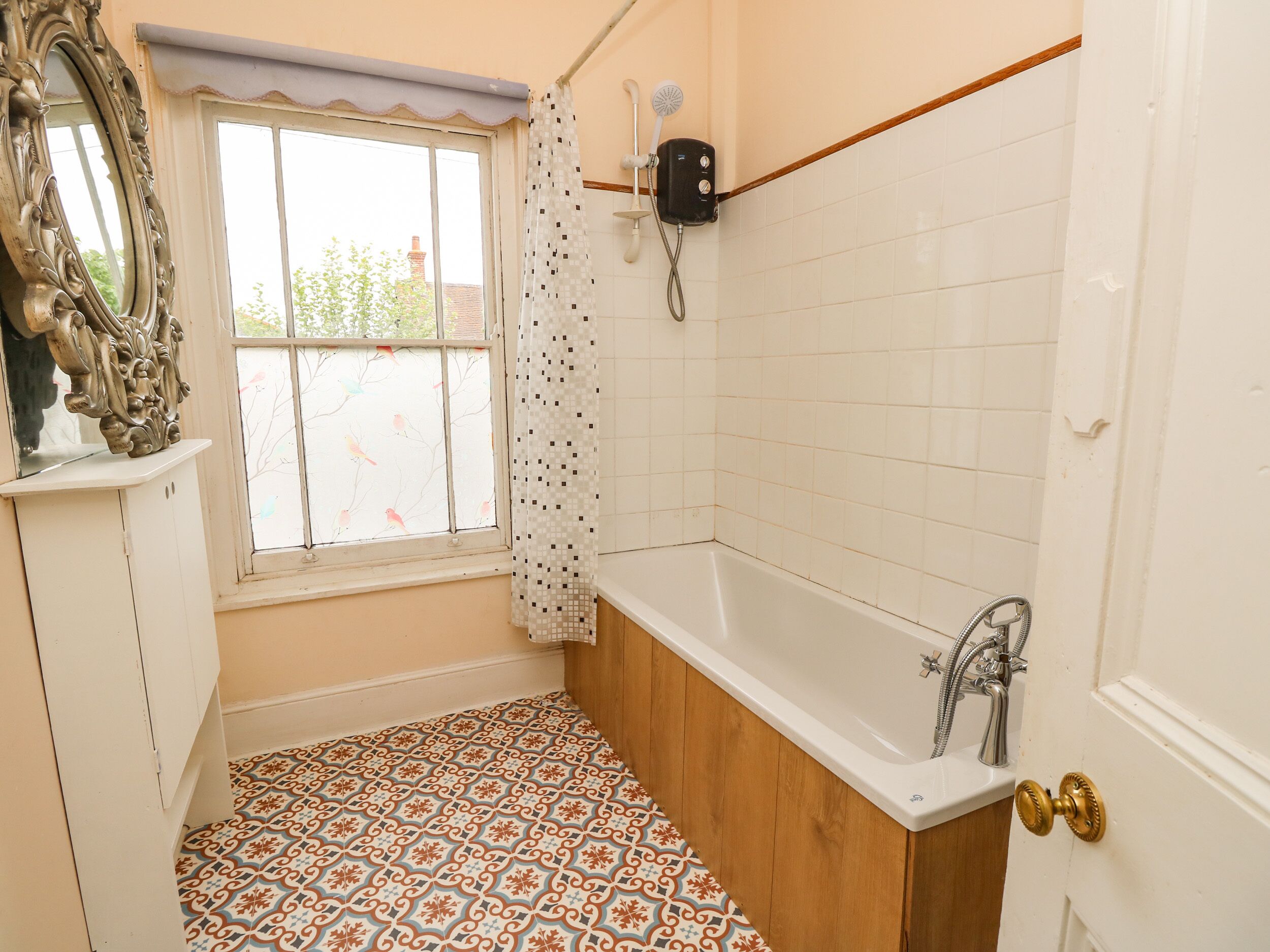 A bathroom with a bathtub, a window with a curtain, a mirror, and patterned floor tiles.