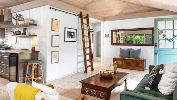 Un vaste espace de vie doté d’un plafond en bois, d’un canapé vert, d’une table basse et d’un coin cuisine aux murs blancs et aux étagères en bois.