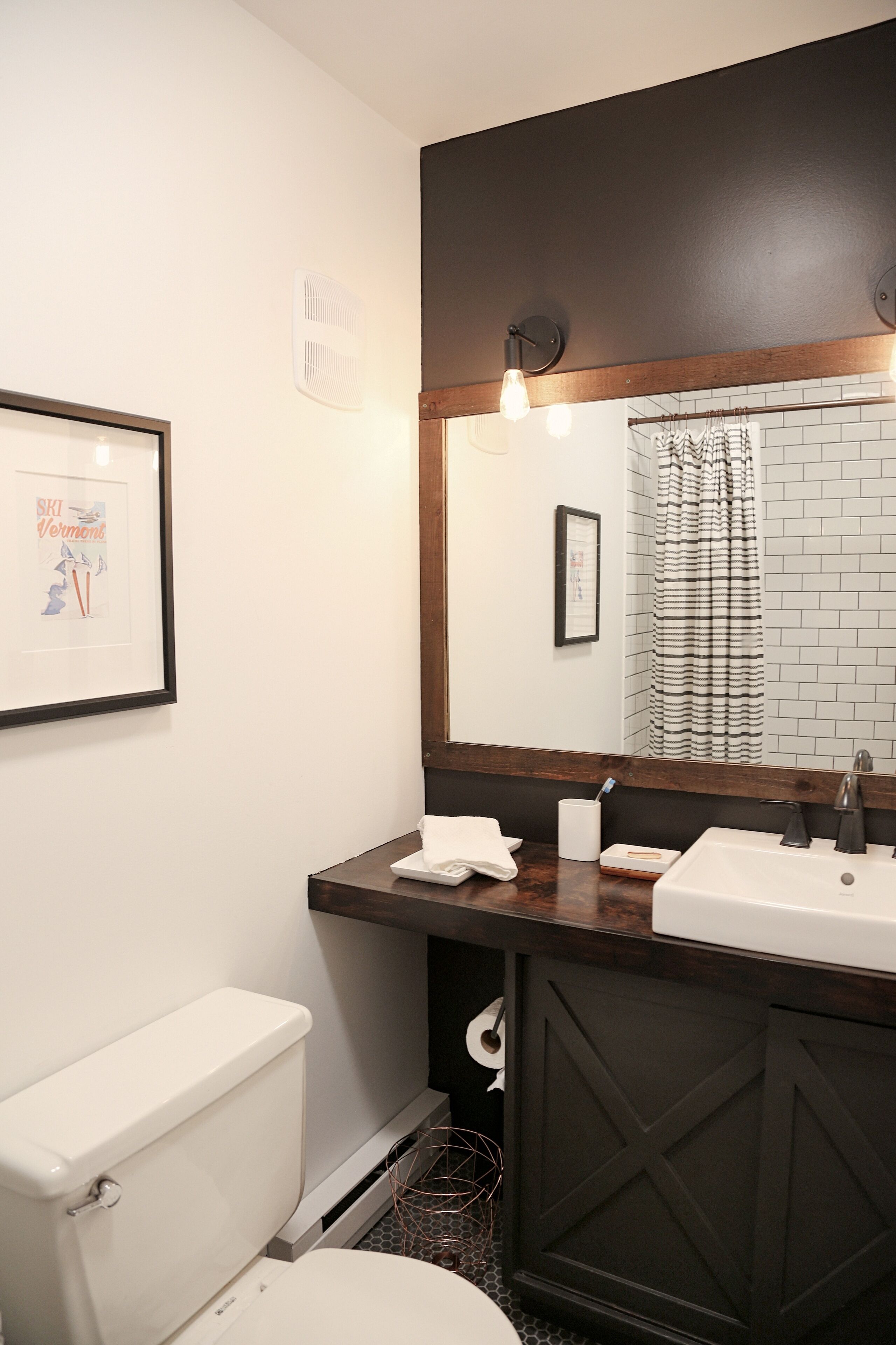 Un baño con inodoro blanco, tocador de madera con espejo, y espejo con marco negro sobre el lavabo.