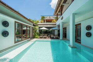 Ein Swimmingpool mit Liegestühlen und Sonnenschirmen, umgeben von einem zweistöckigen Gebäude mit großen Fenstern und einer überdachten Terrasse.