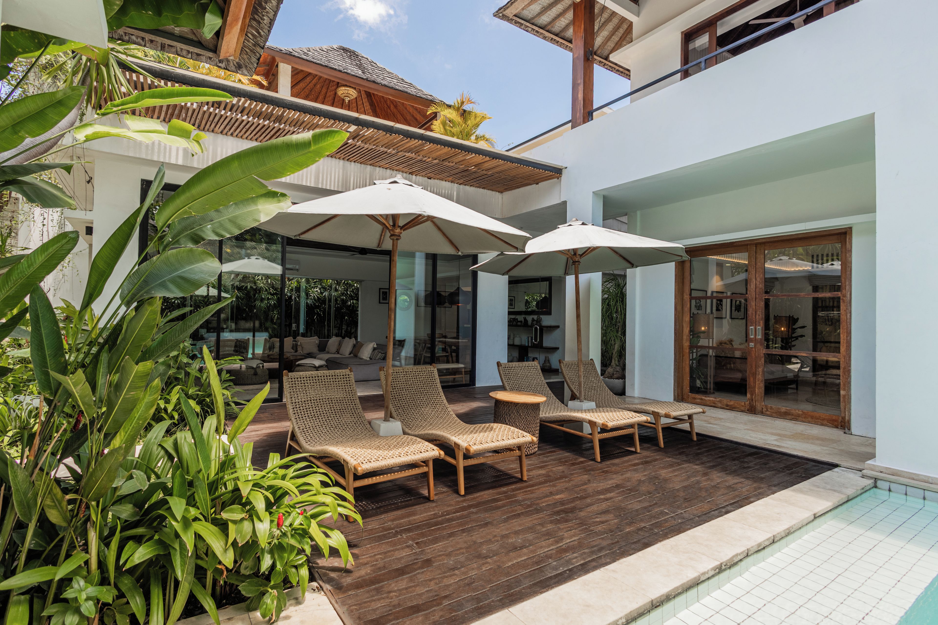 A poolside area with wicker lounge chairs and umbrellas, a wooden deck, and a glass door leading to an indoor space.