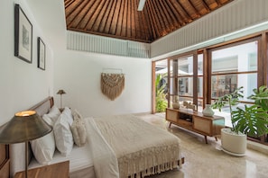 Un dormitorio con cama, mesita de noche, lámpara, decoración en la pared, un colgador de macramé y ventanas amplias con plantas.