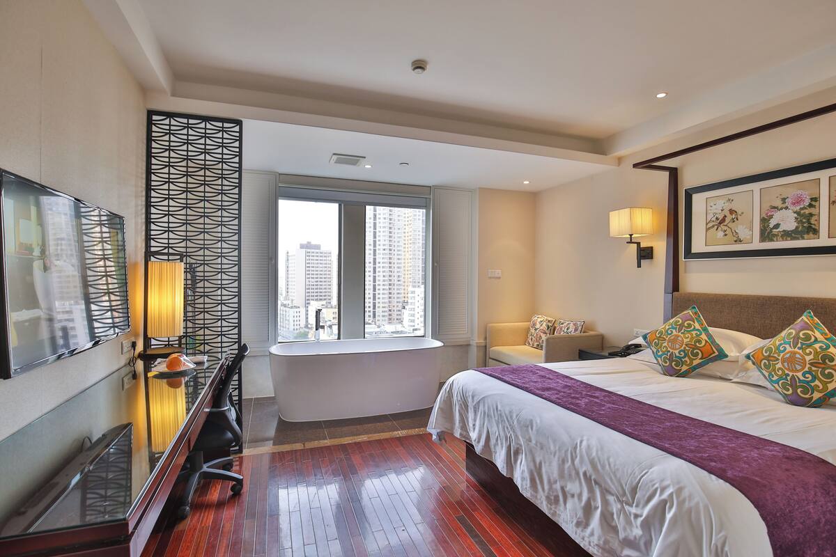 A hotel room with a large bed, a desk with a chair, a window with a view of buildings, a TV mounted on the wall, and a decorative wall panel.