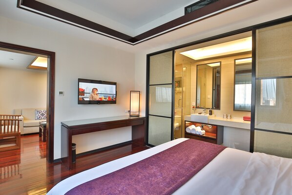 Habitación de hotel con cama, escritorio, televisor y baño con espejo, con vista a otra habitación.