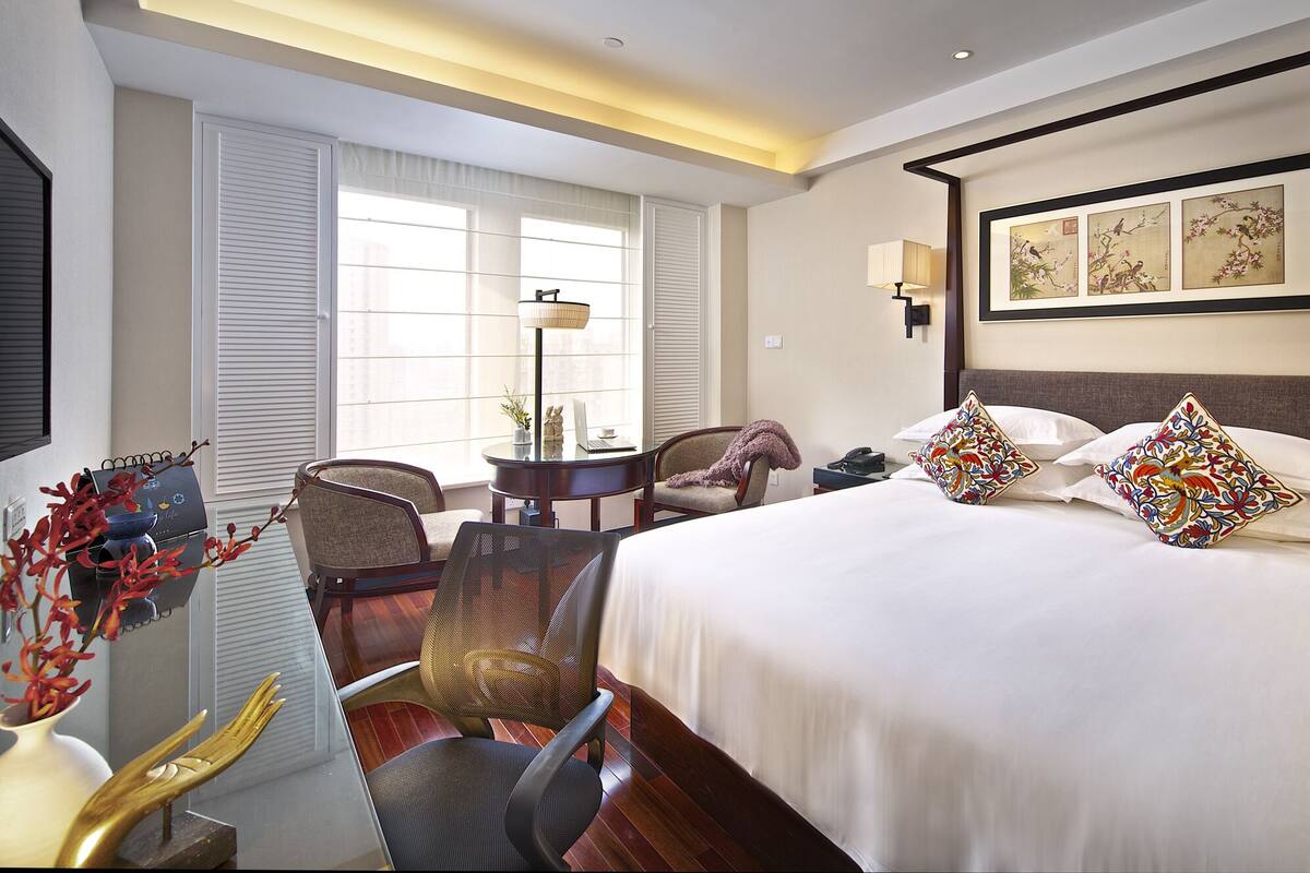 A hotel room with a large bed, two chairs, a desk with a laptop, and a window with blinds.
