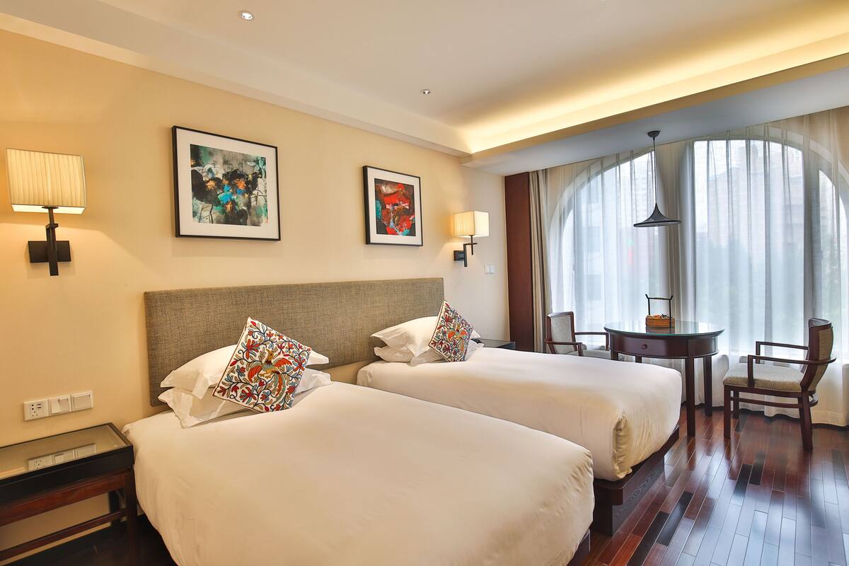 A hotel room with two beds, a desk, a chair, and a large window with curtains.