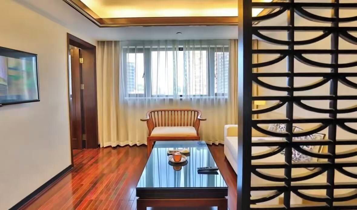 Une chambre d’hôtel avec un plancher en bois, une table basse en verre, un fauteuil rembourré, un canapé, une fenêtre avec des rideaux et un paravent décoratif.