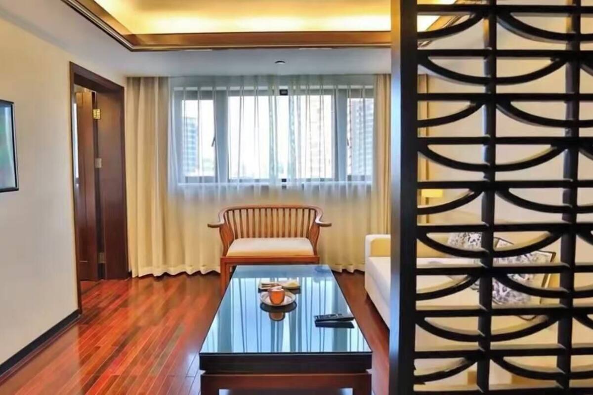 A hotel room with a wooden floor, a glass coffee table, a cushioned chair, a sofa, a window with curtains, and a decorative screen.