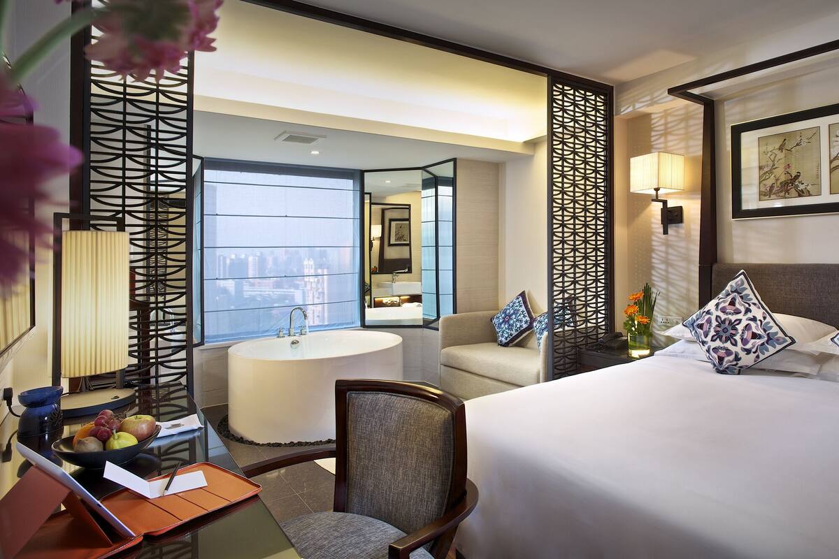 A hotel room with a bed, a desk with a lamp, a chair, a bathroom with a sink, and a view of the city.