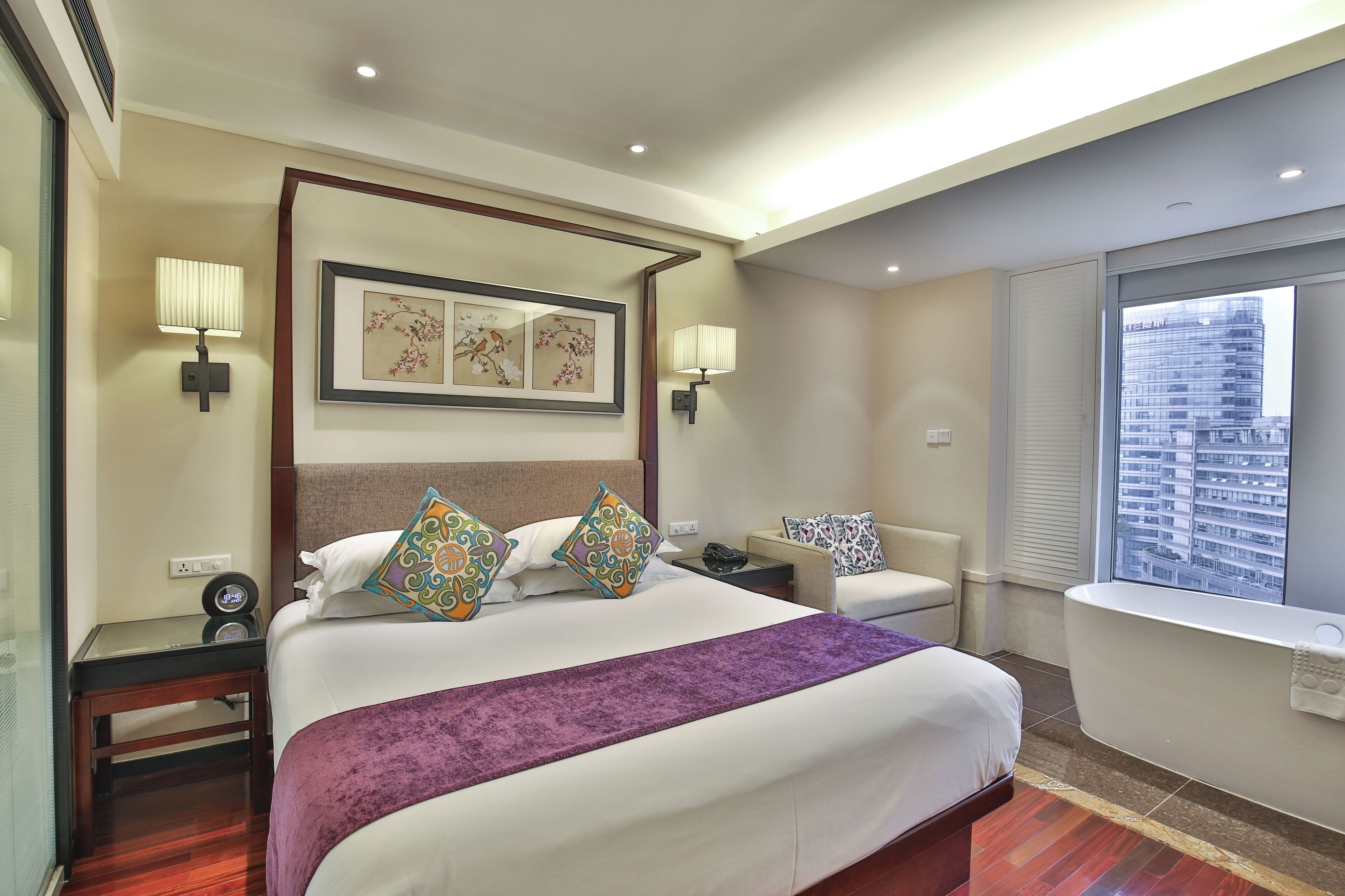A hotel room with a bed, bedside tables, a chair, a window with a city view, and a bathroom with a bathtub.