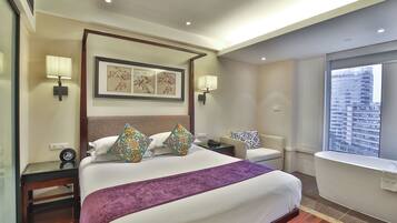 A hotel room with a bed, bedside tables, a chair, a window with a city view, and a bathroom with a bathtub.