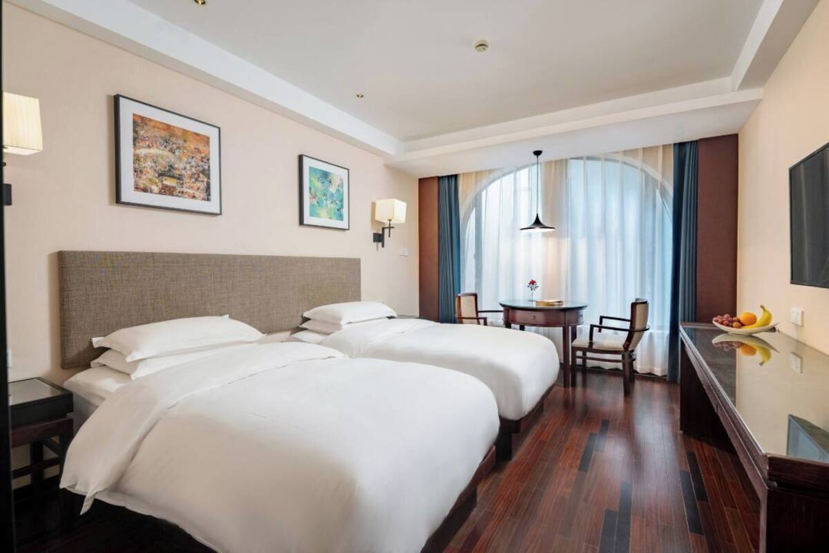 A hotel room with two beds, a desk, and a chair. There are two framed pictures on the wall, a lamp, and a window with curtains.
