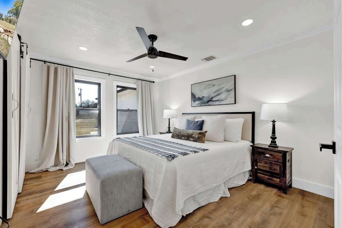 A bedroom with a bed, bedside tables, a ceiling fan, a window with curtains, and a painting on the wall.