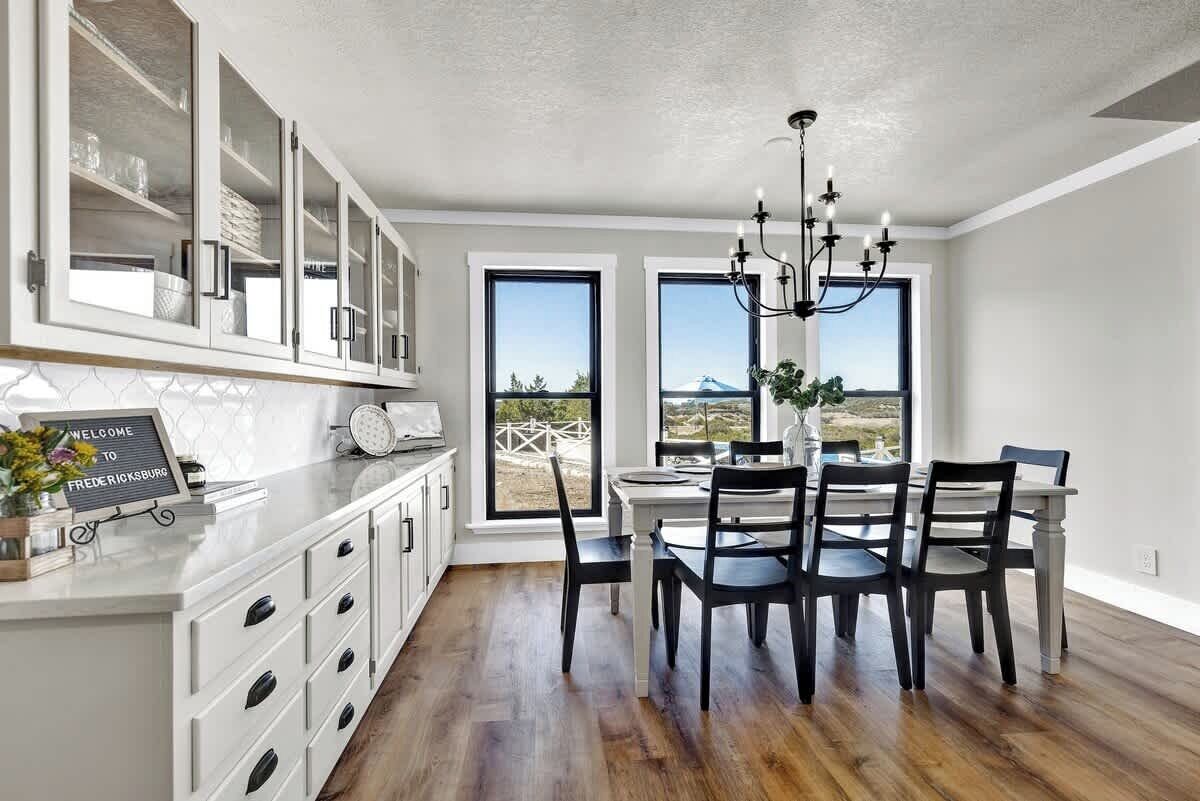 A modern kitchen with white cabinetry, a dining area with black chairs, and large windows offering a view of the outdoors.