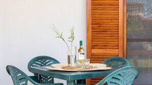 A table with a bottle and glasses, two chairs, a wooden shutter, and a white wall.
