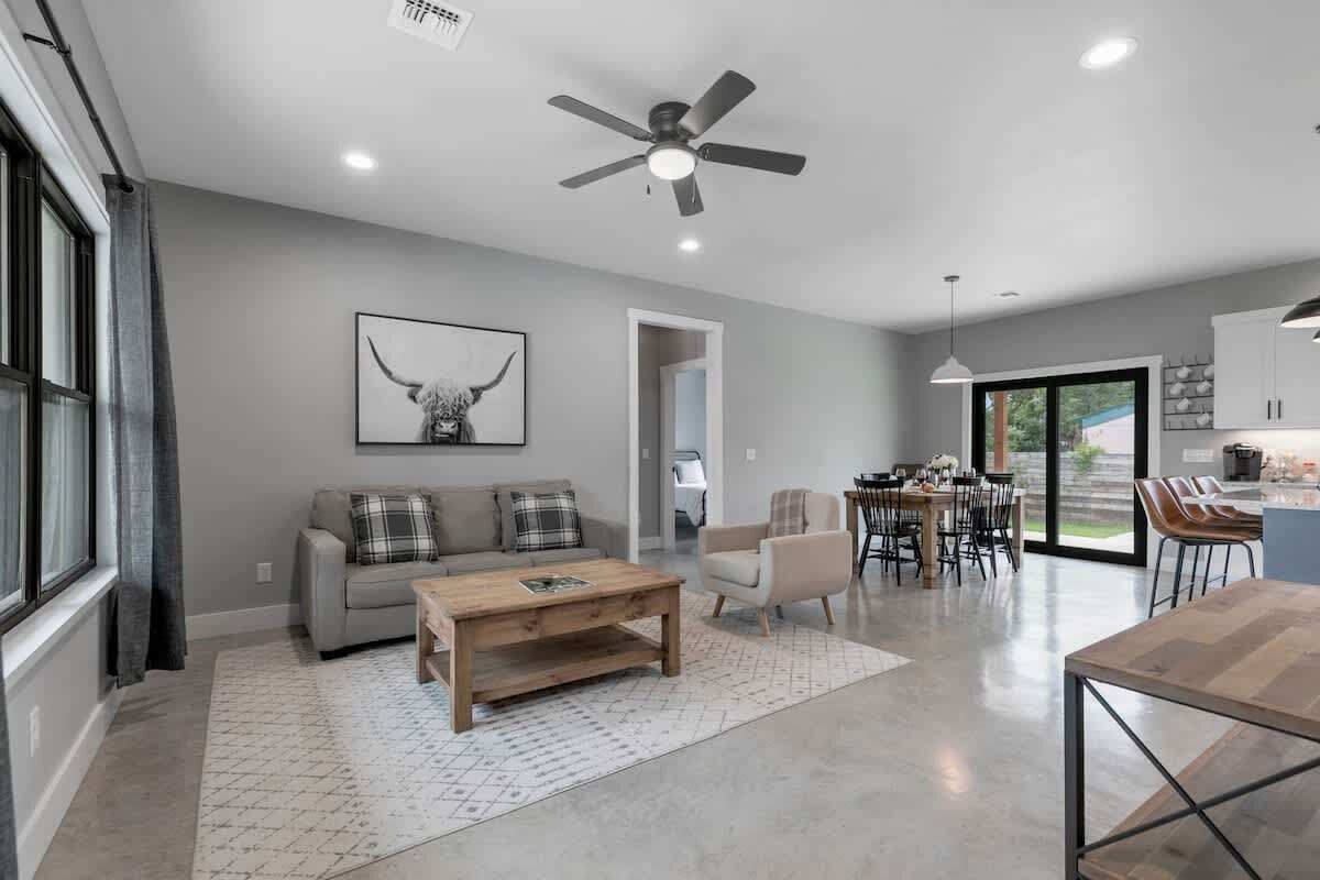 A spacious living area with a gray sofa, a wooden coffee table, and a dining table with chairs. A ceiling fan and recessed lighting are visible.
