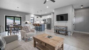A modern kitchen with a central island, a dining area with a table and chairs, a living room with a sofa and coffee table, and a ceiling fan. - Luxury Home 3blck From Main St With Firepit (Fredericksburg)