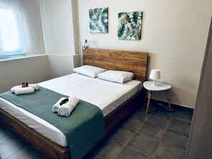 Un lit bien fait, avec du linge de lit blanc, une couverture verte et des serviettes enroulées. Deux œuvres d’art représentant des feuilles tropicales sont accrochées au mur. Une petite table blanche avec une lampe se trouve à côté du lit.