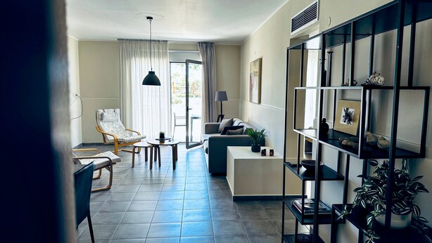 Uma sala de estar moderna com sofá, poltrona e mesa de centro, uma área de jantar com cadeiras e uma estante com objetos decorativos.