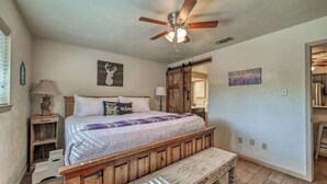 Une chambre à coucher avec un lit en bois, un banc, un ventilateur de plafond et une porte donnant sur une autre pièce.