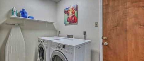 Una sala de lavandería con dos lavadoras blancas, un estante con productos de limpieza y un cuadro en la pared.