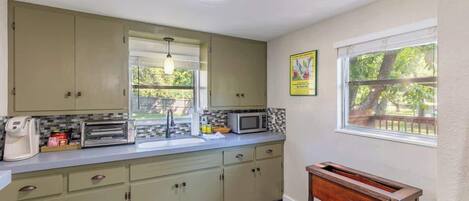 Una cocina con armarios verde claro, un microondas, un tostador y una ventana con vistas a árboles.