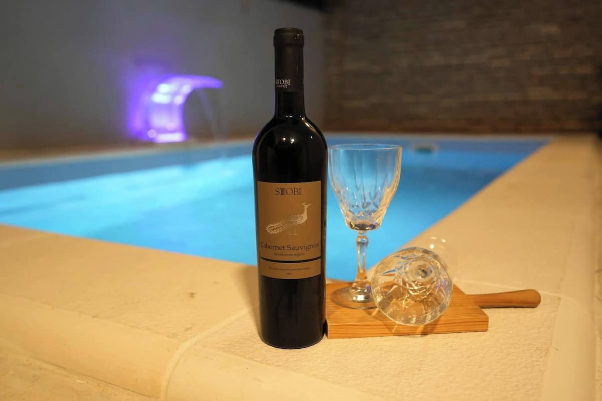 A bottle of Stobi Cabernet Sauvignon wine, a wine glass, and a wooden cutting board with a knife on a table near a pool.