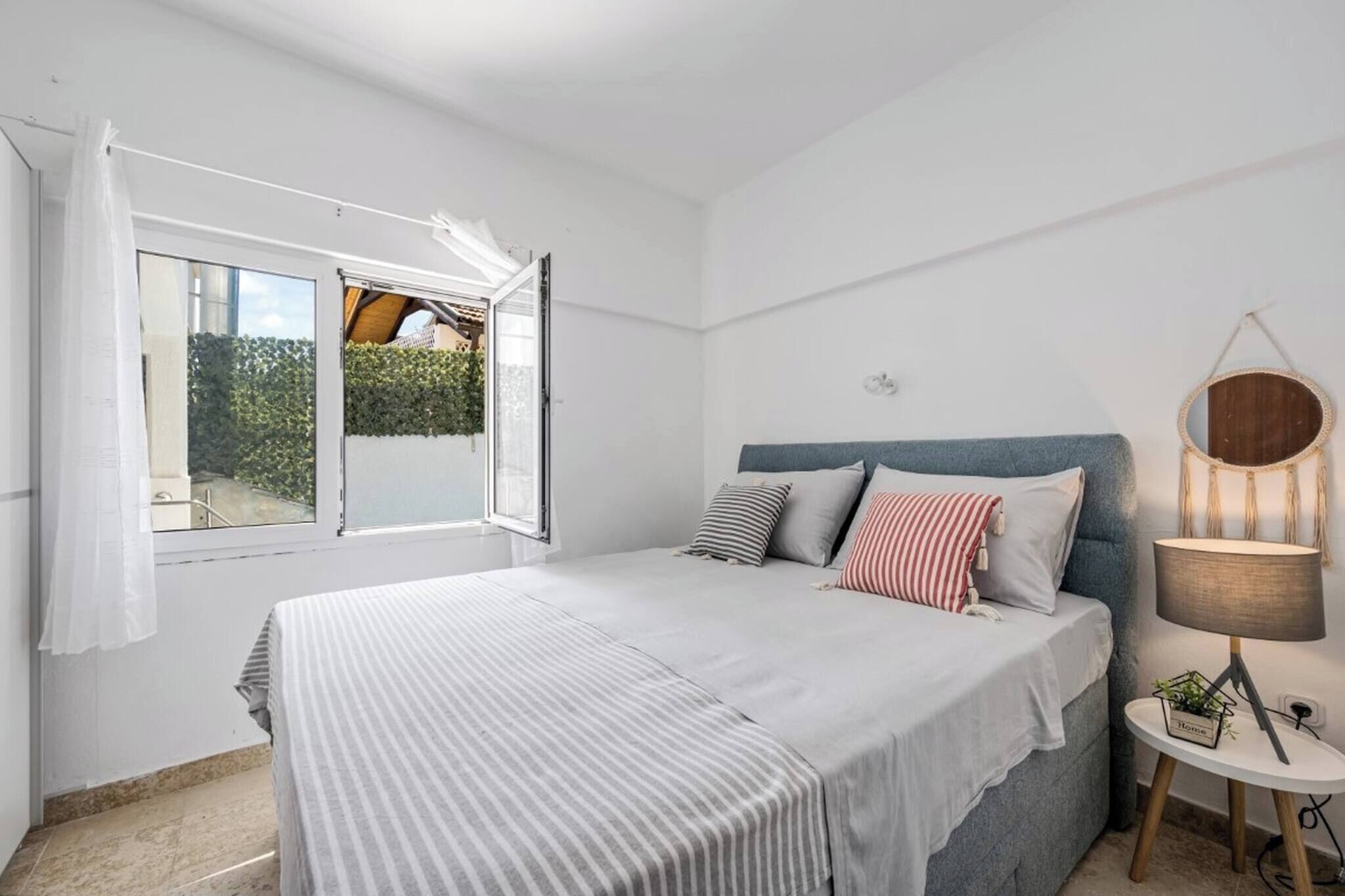 Quarto com cama, mesa de cabeceira com abajur, janela com cortinas e vista para o verde lá fora.