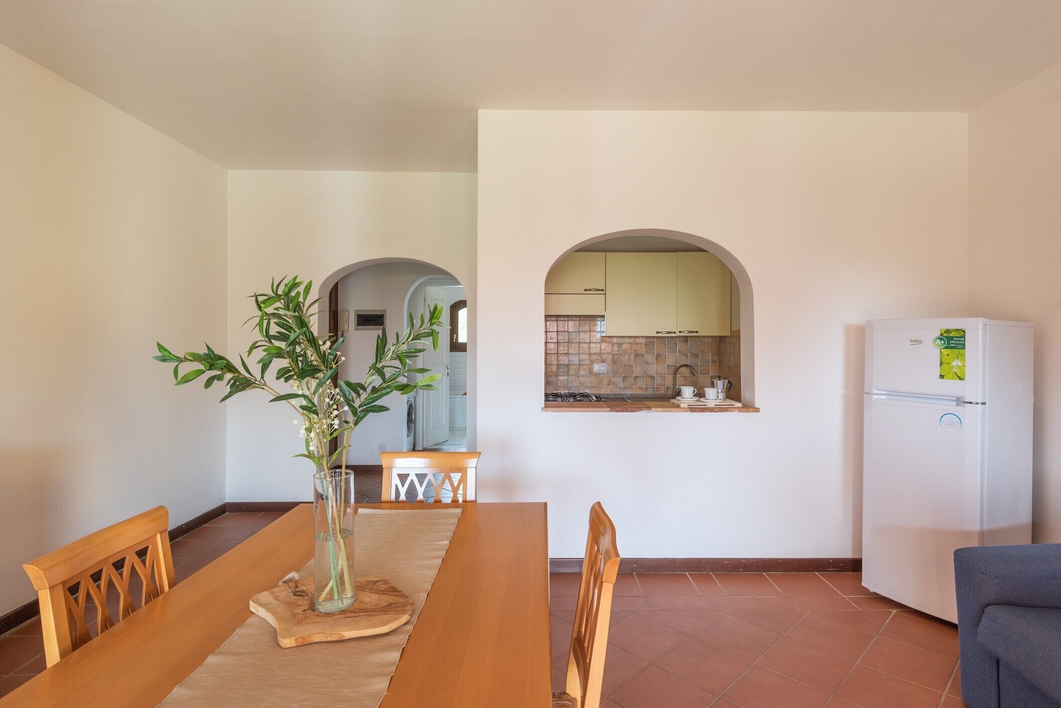Un coin repas avec une table en bois, des chaises et un vase contenant des plantes vertes. On aperçoit une cuisine à travers une arche en arrière-plan.